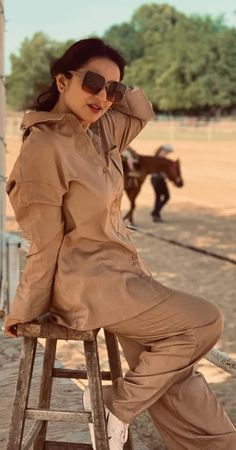 a woman sitting on top of a wooden stool in front of a horse pen wearing sunglasses