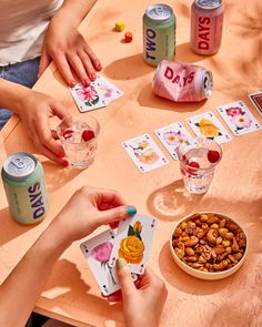 two people playing cards at a table with drinks and snacks on it while another person holds up their hand