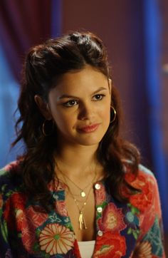 a woman with long dark hair wearing a colorful shirt and gold hoop earrings is looking at the camera