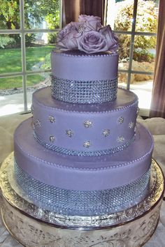 a three tiered cake with purple frosting and flowers on top is displayed in front of a window