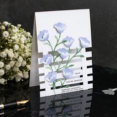 a white card with blue flowers on it next to a pen and flower arrangement in the background