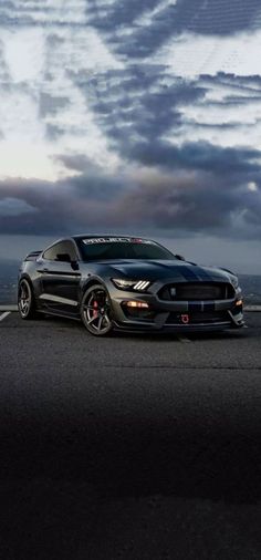 a black mustang parked on the side of a road under cloudy skies with clouds in the background