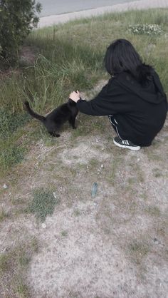 a woman kneeling down next to a black cat on top of a grass covered field