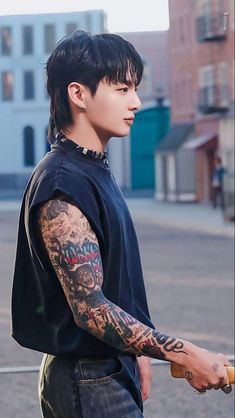 a man with black hair and tattoos on his arm holding a skateboard in the street