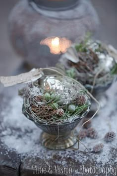 two silver bowls filled with plants and pine cones on top of a table next to a candle