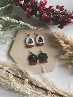 three small christmas tree earrings sitting on top of a table next to some dried plants