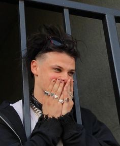 a man with black hair and piercings covers his mouth as he looks out the window