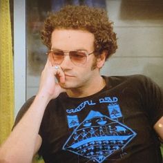 a man with curly hair and sunglasses talking on a cell phone while sitting in front of a window