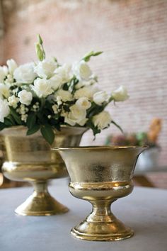 two gold vases filled with white flowers on top of a table next to each other