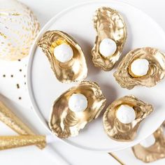 four gold oysters on a white plate next to confetti sticks