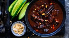 a bowl of stew next to cucumbers and spoons on a black surface