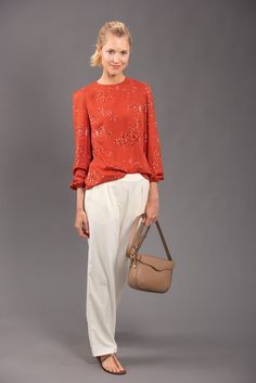 a woman in an orange top and white pants holding a handbag while standing on a gray background