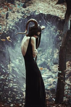 a woman in a black dress standing next to a tree with horns on her head