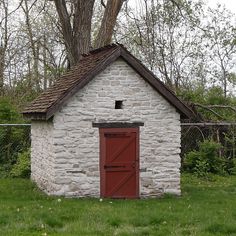 a small brick building with a red door in the grass next to a fence and trees