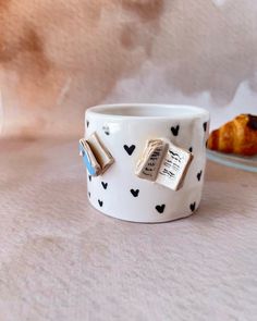 a white cup with black hearts and books on it sitting on a table next to a muffin