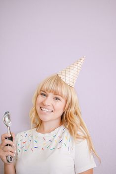 a woman wearing a party hat holding a spoon and looking at the camera while standing in front of a purple wall