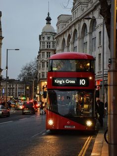 a red double decker bus driving down a street next to tall buildings and traffic lights