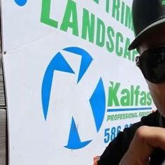 a man wearing sunglasses and a hat holding a cell phone to his ear while standing in front of a sign