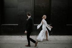 a bride and groom walking down the street in front of a black building with tall windows