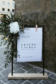 a sign with flowers on it sitting in front of a stone wall next to a plant