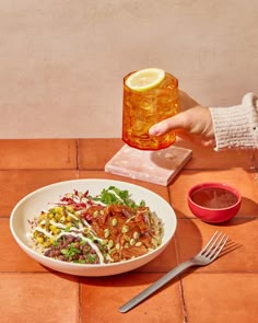 a person is holding a drink over a bowl of food on a table with utensils