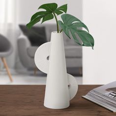 a white vase with a green plant in it on a table next to a magazine