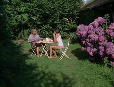 two women sitting at a table outside in the grass with purple flowers behind them, eating cake