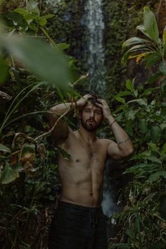 a shirtless man standing in front of a waterfall with his hands on his head