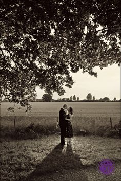 a man and woman standing under a tree in the middle of a field with their arms around each other