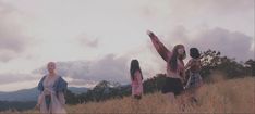 three women are walking in a field with clouds behind them and one woman is wearing a pink dress