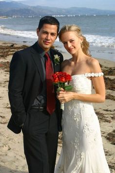 a man in a suit and tie standing next to a woman wearing a wedding dress on the beach