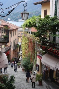 people are walking down an alley way in the mountainside town with flowers growing on the balconies