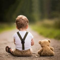 a little boy sitting on the ground next to a teddy bear and wearing suspenders