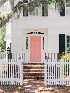 a pink door sits in front of a white house with black shutters and trees