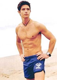 a shirtless man standing on the beach with his hands in his pockets