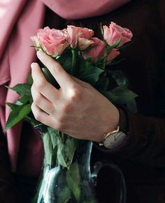 a woman holding pink roses in a clear vase with green leaves on the bottom and one hand