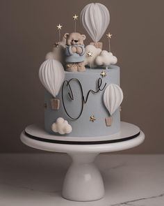 a blue cake decorated with hot air balloons and a teddy bear on top is sitting on a pedestal