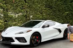 a man kneeling down next to a white sports car in front of some trees and bushes