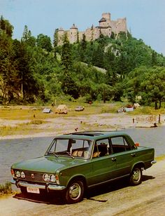 an old green car is parked in front of a lake with a castle on top