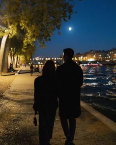 two people are walking down the sidewalk by the water at night, one is holding an umbrella