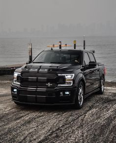 a black truck parked on top of a gravel road next to the ocean in front of a city skyline