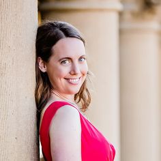 a woman in a red dress leans against a column and smiles at the camera with her eyes closed