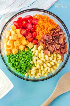 a bowl filled with pasta, ham, cheese and peas next to a wooden spoon