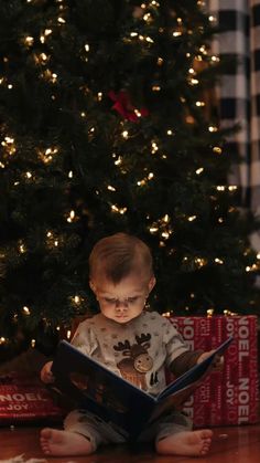 a baby sitting in front of a christmas tree while reading a book with the lights on