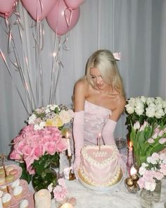 a woman sitting at a table with a cake and flowers in front of her, surrounded by pink balloons