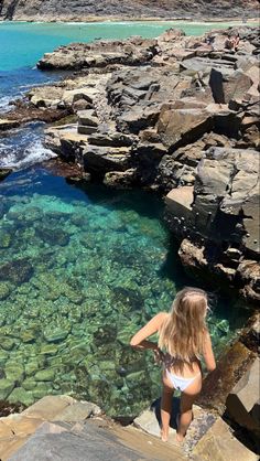 a woman standing on some rocks near the ocean with clear blue water in front of her