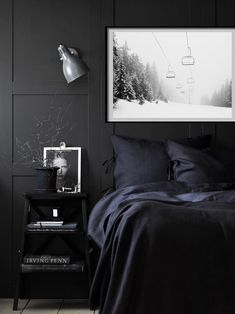 a bed with black sheets and pillows in a dark room next to a lamp on the wall