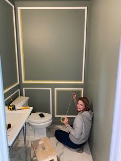 a woman sitting on the floor in front of a toilet and painting walls with yellow tape