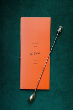 an orange book sitting on top of a green table next to a silver spoon with the word f drink written on it