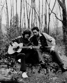 two people sitting on a log in the woods, one playing a guitar and the other singing